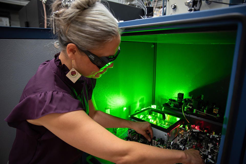 A researcher wearing safety glasses reaches into a box of circuitry and other equipment, which emits a green glow.