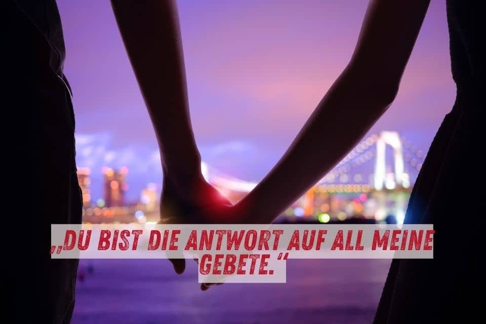 Das Bild zeigt zwei Personen, die sich bei Sonnenuntergang an den Händen halten. Im Hintergrund sind Lichter einer Stadt und eine beleuchtete Brücke zu sehen. Unten im Bild steht der Text: