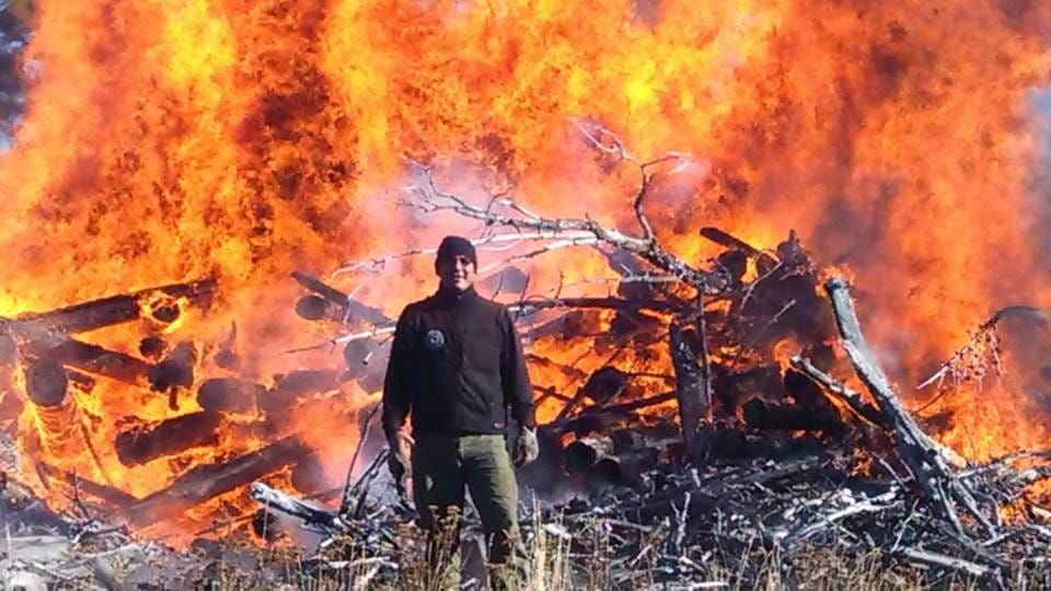 Representative Tyson Runningwolf stands in front of a large wildfire.