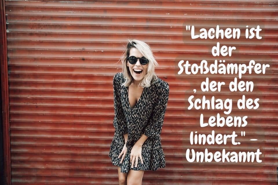 Das Bild zeigt eine lächelnde Frau in einem dunklen Kleid mit Sonnenbrille vor einer roten Wellblechwand. Rechts steht der Text: