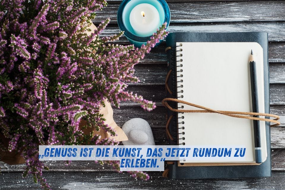 Das Bild zeigt ein Notizbuch mit einem Stift darauf, daneben eine blaue brennende Kerze und lila Heidekraut auf einem Holztisch. Darunter steht der Text: „Genuss ist die Kunst, das Jetzt rundum zu erleben.“