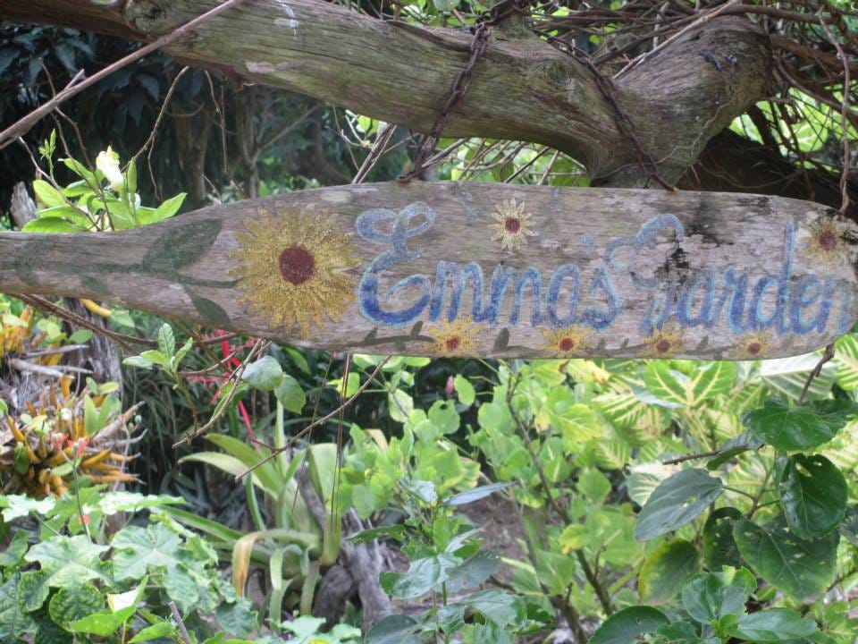 Hand painted woold sign with a big sunflower and the words Emma’s Garden. Tropical plants grow below the sign.