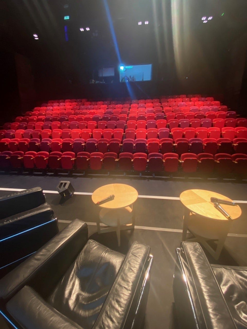 An empty theatre. Rows of red seats waiting to be folded down and sat on. Two small round tables on the stage with microphones on them and black chairs. The stage is set.