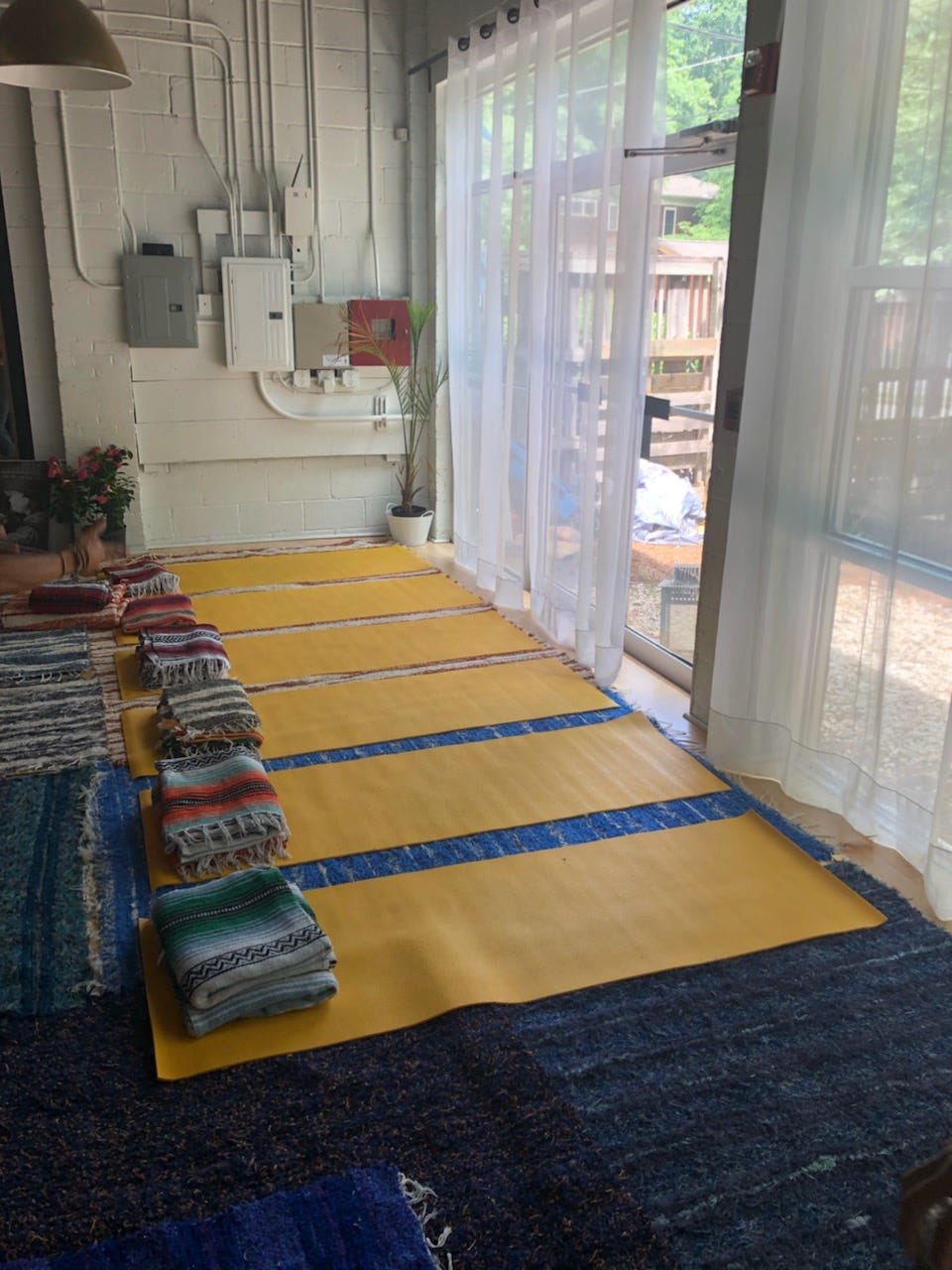 Yellow mats and blankets line the floors of The Yellow Mat Yoga Studio. An open, partially curtained window lets in a soft breeze.