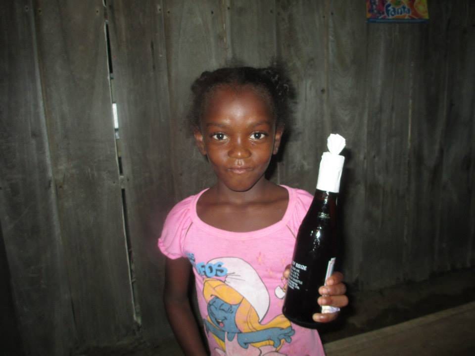 Petite black young girl with wide and a cheeky closed mouth smile holds a bottle of beer. She’s wearing a pink t-shirt with Smurfette on the front.