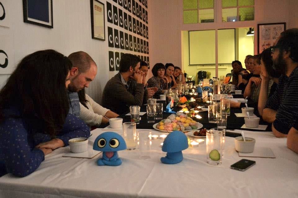 a group of people sit around a long table, facing towards a man speaking who is seated in the middle. The table is laden with tealights, character toys, empty gin glasses and plates stacked high with multi-coloured mochi. White ensos on black paper adorn the walls.