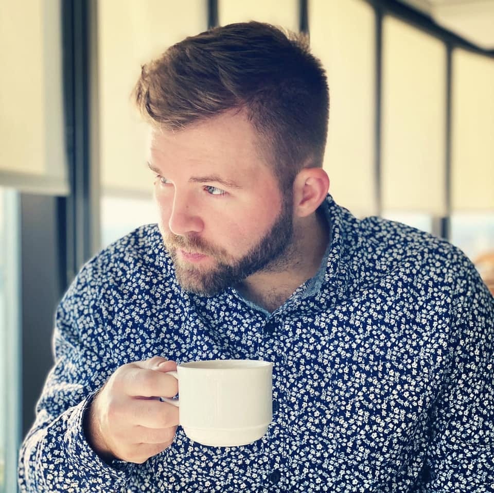A bearded man in a blue patterned shirt holds a coffee cup and looks out of the window.