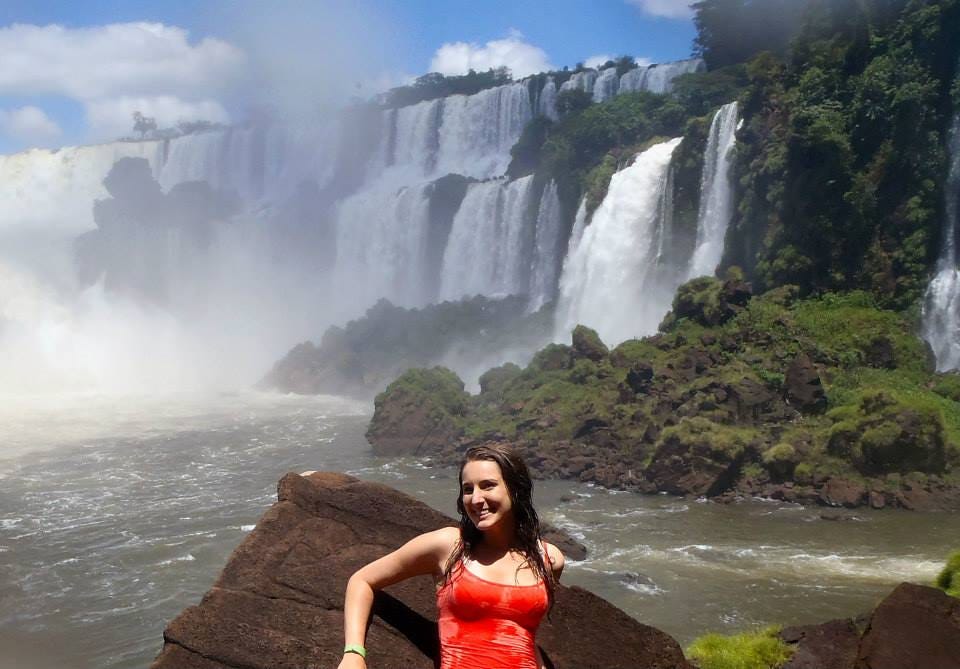 A girl in a pink tank top poses in front of multiple layers of waterfalls surrounded by lush rainforest at Iguazu Falls.
