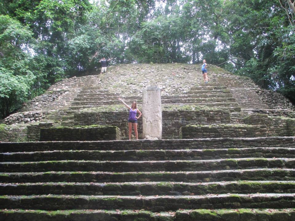 Mayan temple structure with stairs that has an inset level from the first and the three children are dispersed standing on the stairs from the middle of the structure up to the top.