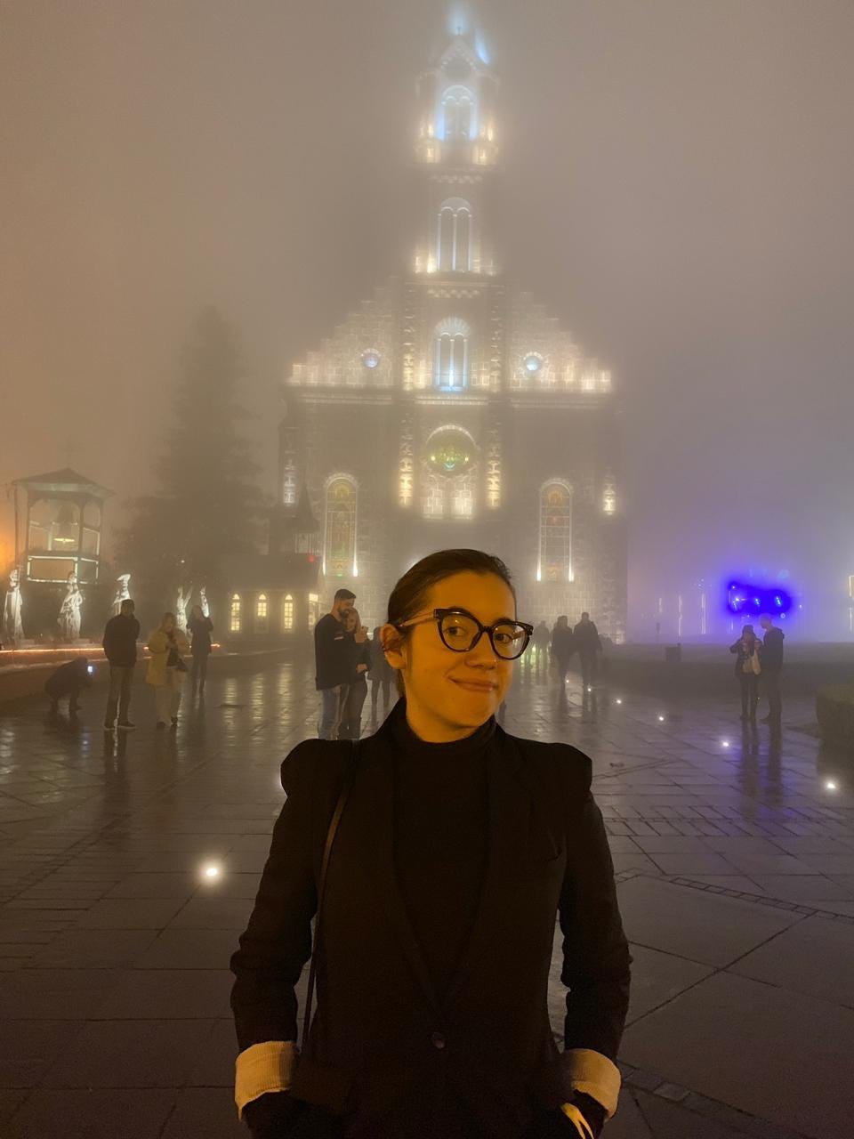 Primeiro plano: eu de cabelo preso, blazer preto sobre suéter preto e óculos. Fundo: igreja coberta por muita névoa.