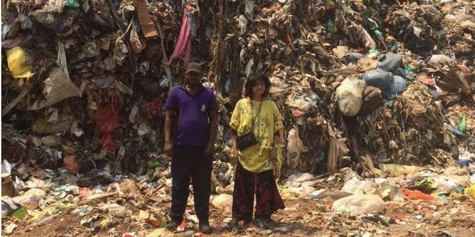 Two people stand in front of a textile rubbish heap which towers above them