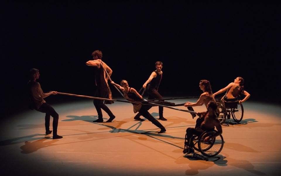 Group dancing onstage connecting with each other by rods where three dancers are in wheelchairs and four dancers are standing.