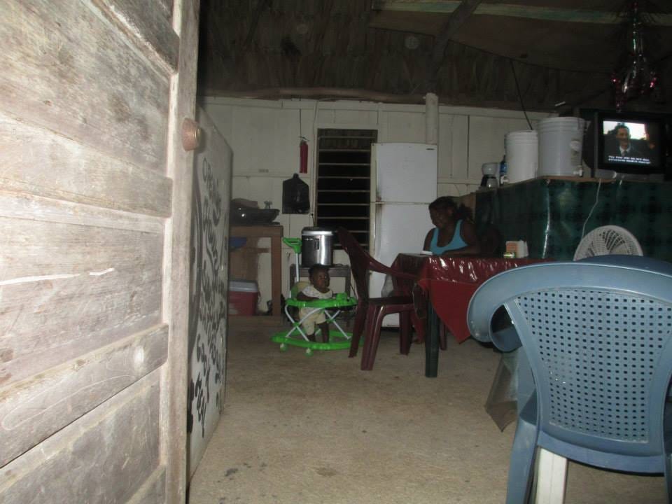A woman sits behind a plastic square table next to a baby in a walker in a room that resembles an open garage with a makeshift kitchen.