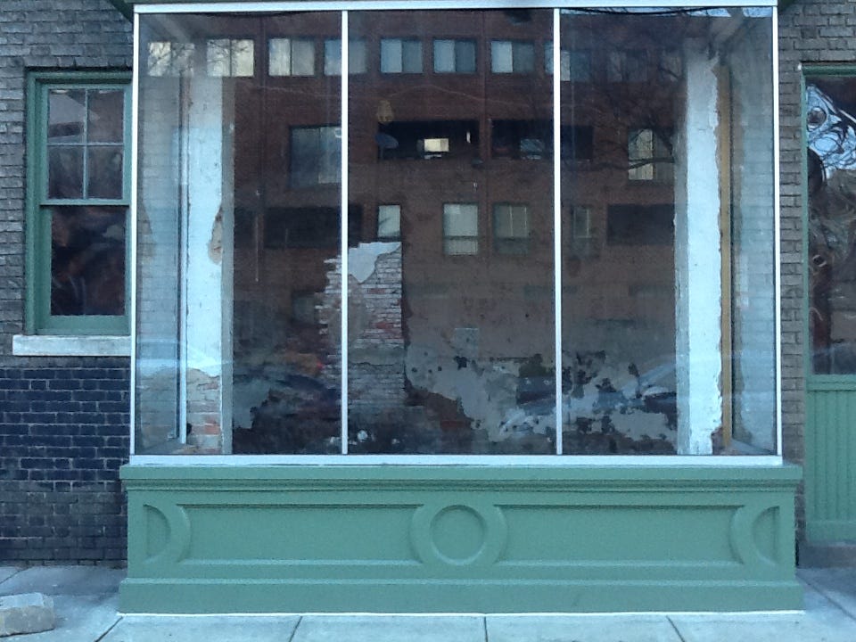 Photograph of a glass picture-window of a storefront, in which the glass reveals a view of demolition happening inside the storefront, while it also reflects the image of the exterior of a building across the street. To the left and right side of this large picture-window are smaller windows. The window to the left reflects or reveals nothing, while the window to the right reflects some light.