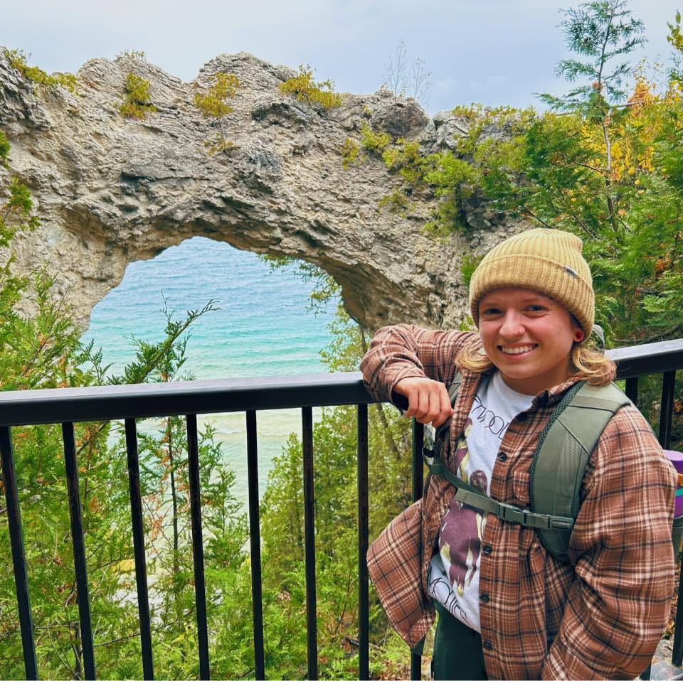 Chapin, a white person with Dwarfism, is smiling. Chapin stands with their arm resting on a guard rail that comes up to their chin. They are standing in front of Pictured Rocks, a natural arch formation of rock that stands in front of bright blue waters.