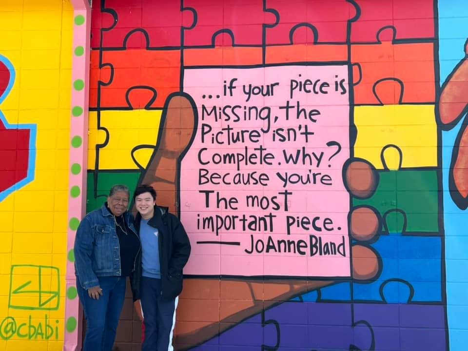 An older Black woman standing next to an Asian college student in front of a mural on a brick wall. The mural is in bright colors and shows a large hand holding a card with a quote.