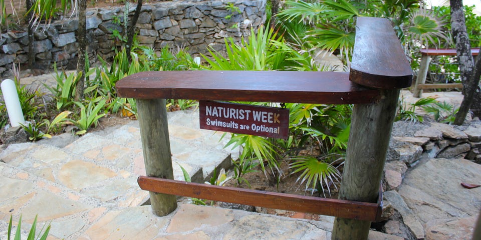 Handmade sign reading “NATURIST WEEK Swimsuits are Optional” attached to a wooden signpost