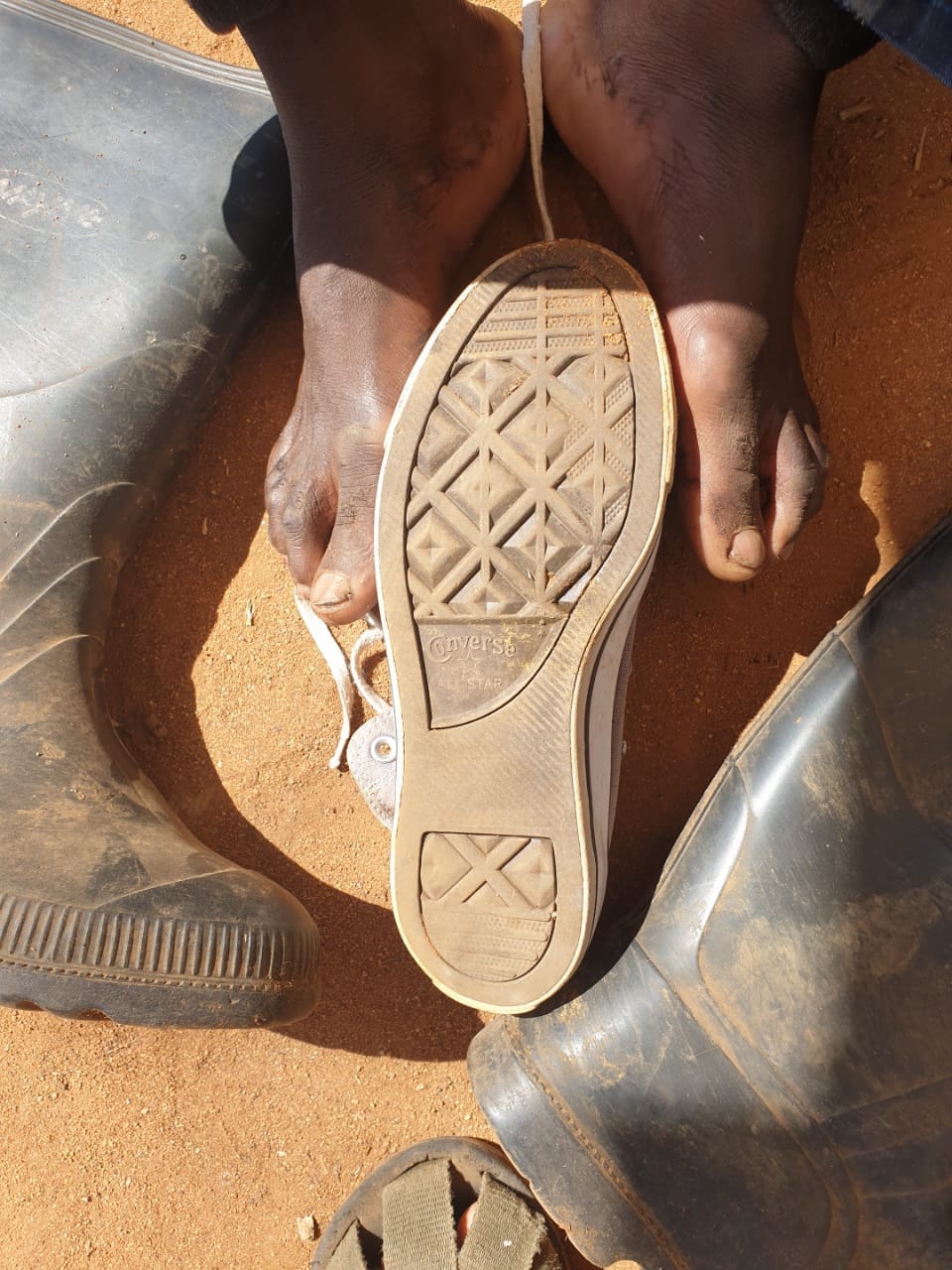 African male feet, a converse shoe and gumboots with soil in the background.