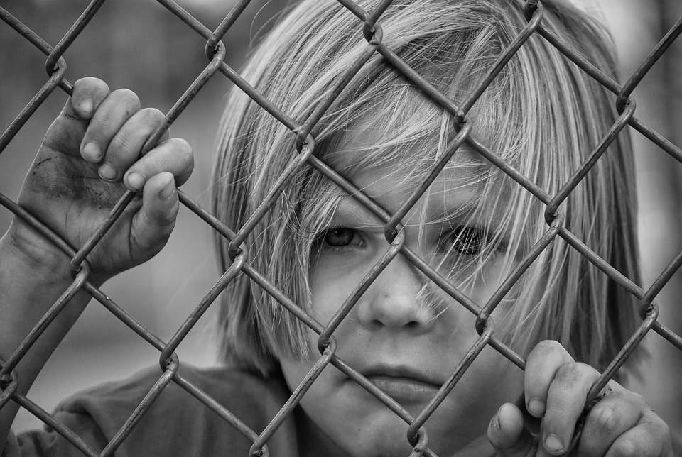 sad child looking out from behind wire fence