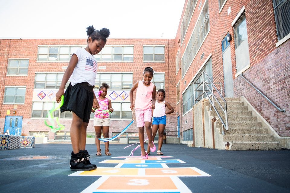 Four elementary school girls in colorful summer clothing play hopscotch with a school building in the background.