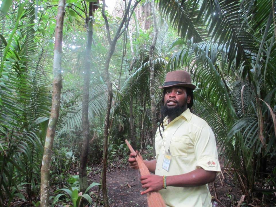 A medium build black man (the jungle guide) stands facing the camera in the jungle. He’s wearing a fedora hat and is carrying a homemade fly-swatter made of grass.