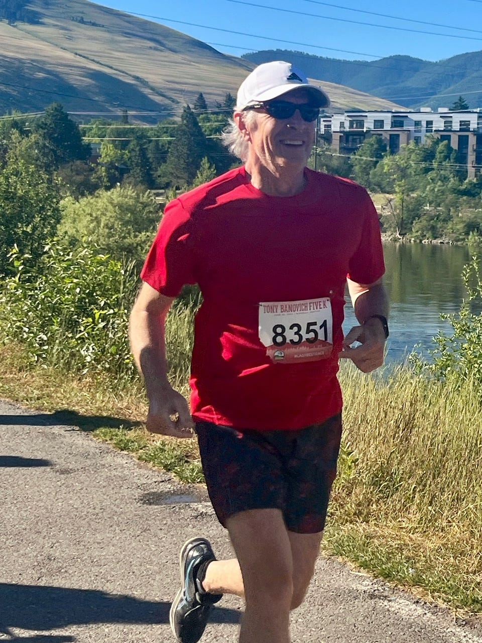 A man with gray hair and a red shirt with a racing bib smiles while running