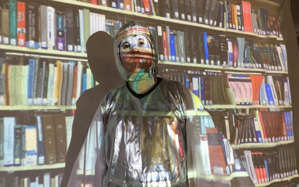 A boy dressed as a superhero looking determined, standing in front of a projection of a bookcase