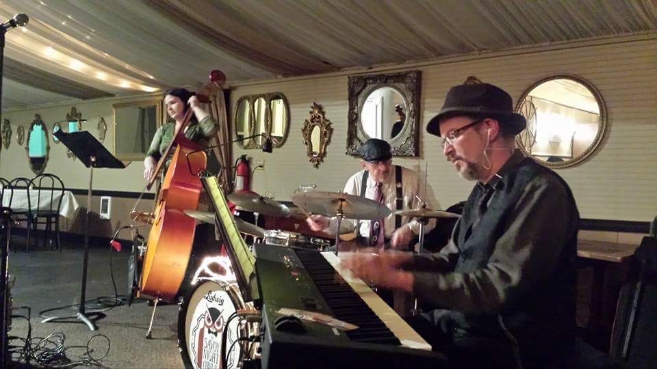 A man sits in the foreground playing piano, wearing a fedora and glasses. Another man is in the background, wearing suspenders and playing the drums. To the left of both men, a woman in a green shirt is standing up playing the stand-up bass.