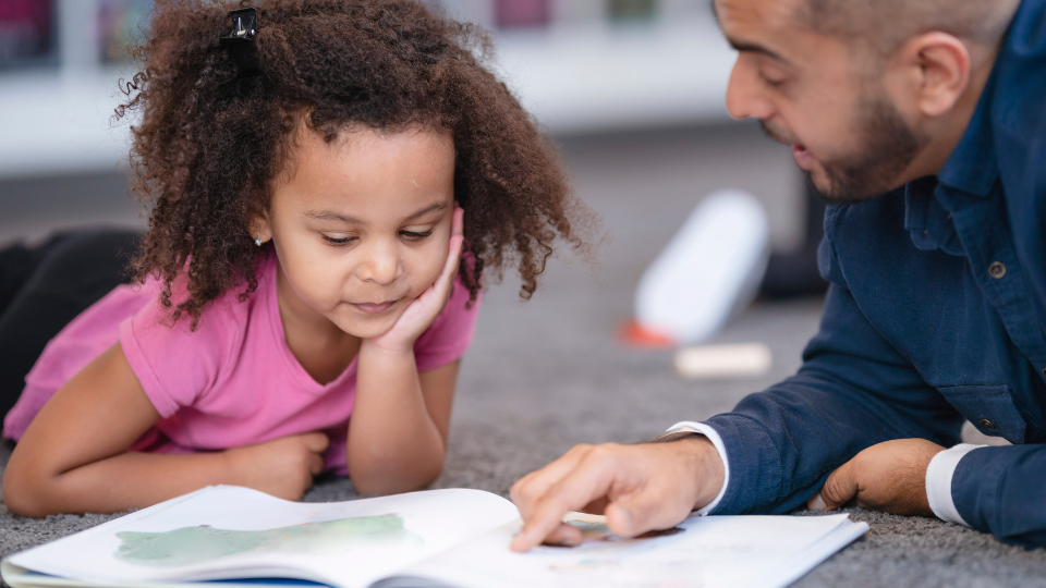 A young girl sits with an adult, reading an open book.