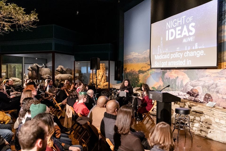 A crowded room with a screen in the front. Three panelists with disabilities sit at the front.