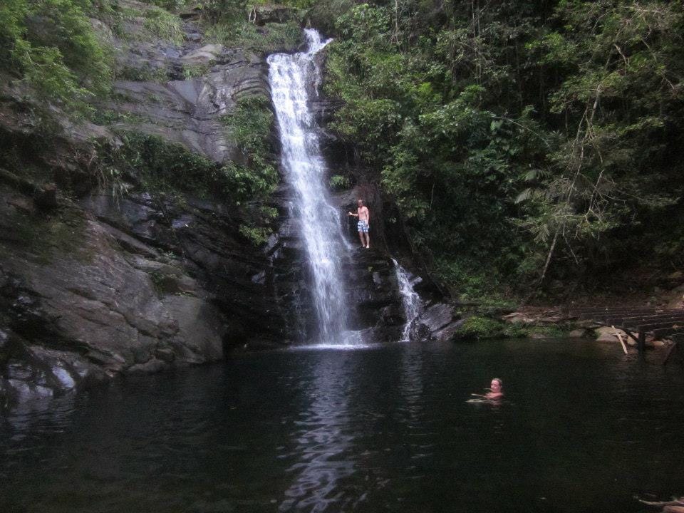 Author’s son stands on a ridge by a waterfall about twenty feet above the pool. The author is in the pool below the waterfall.