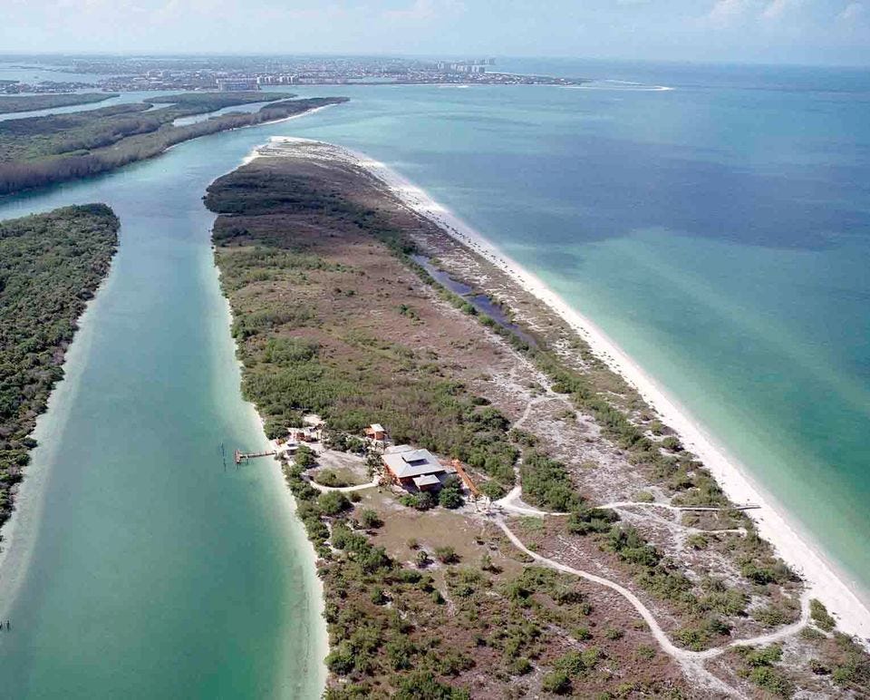 Aerial image of a home on a low sandbar surrounded by blue-green water, with a city skyline visible on the horizon.