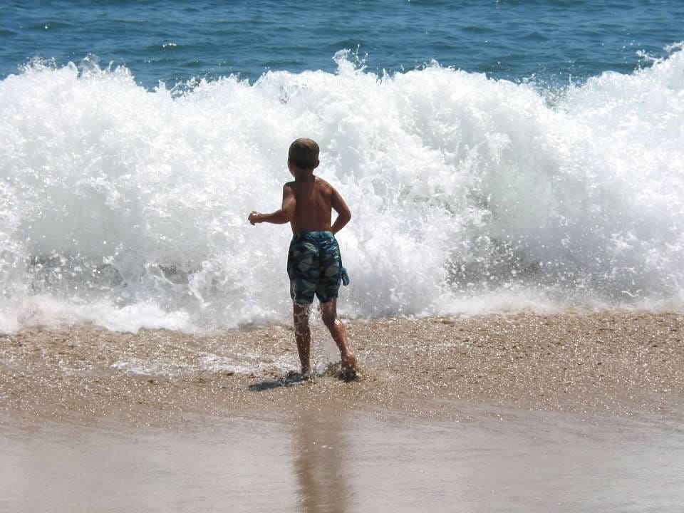 Young boy faces ocean surf higher than his head.