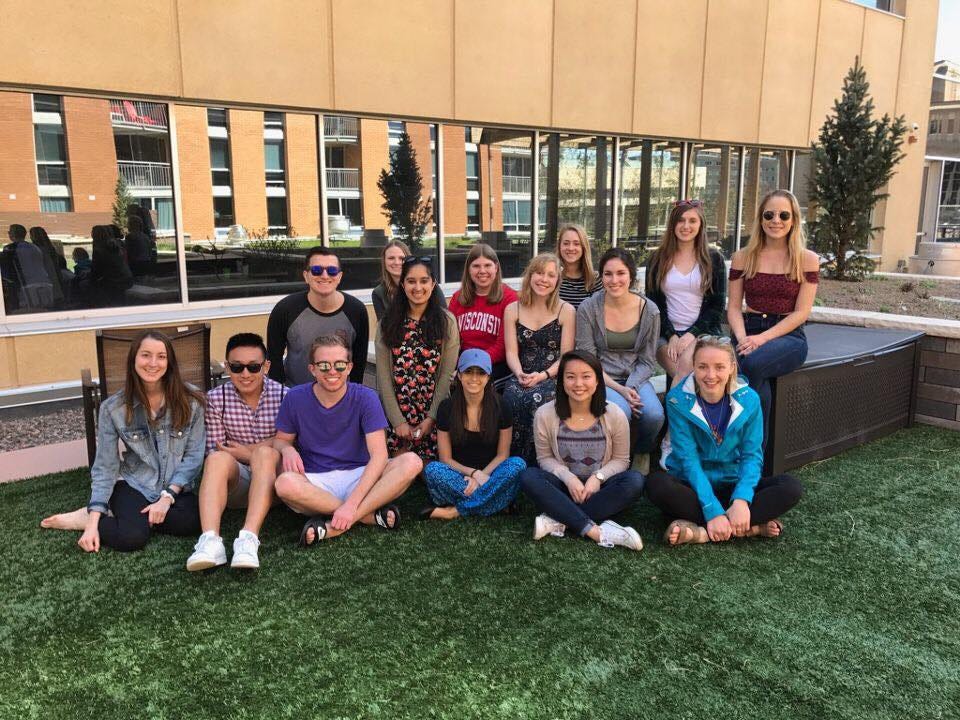 A diverse group of college student pose for a group photo, sitting together on a patch of grass outside.