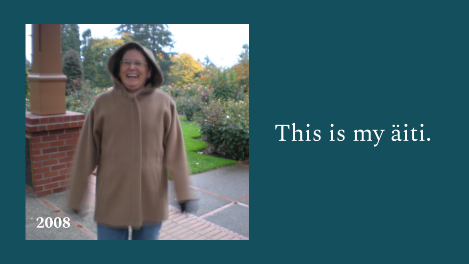 This is my äiti, 2018. With image of a smiling woman with glasses jumping with joy in a hooded brown jacket during a rainy day at the Portland Rose Garden.