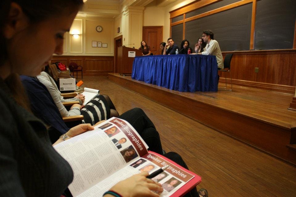 Students in chairs examine event brochures as speakers at a table onstage in front of them deliver a talk.