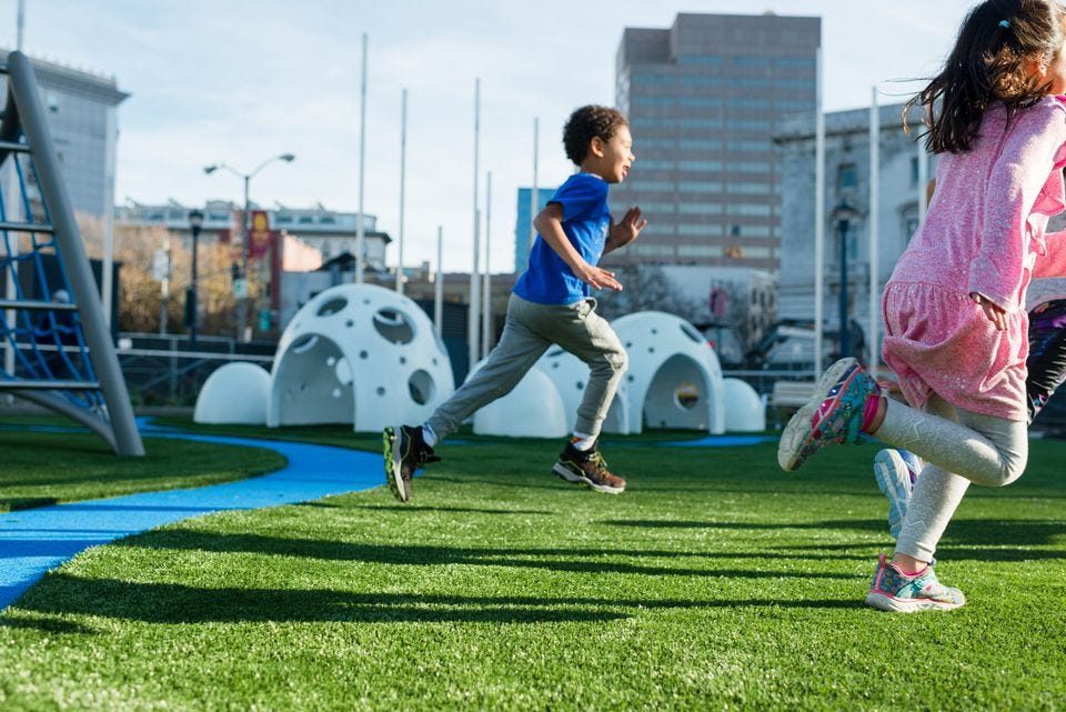 A grade school boy and girl run on a green playground with buildings in the background