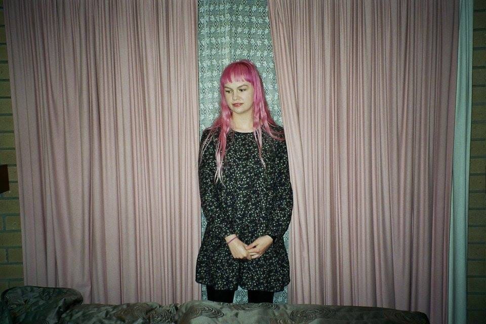 A girl with long wavy bright pink hair and bangs standing in a retro motel room in front of pale pink curtains and lace drapes. She wears a long sleeve flannelette dress with a dainty floral print. She is gazing to her right. The image is taken on 35mm film.