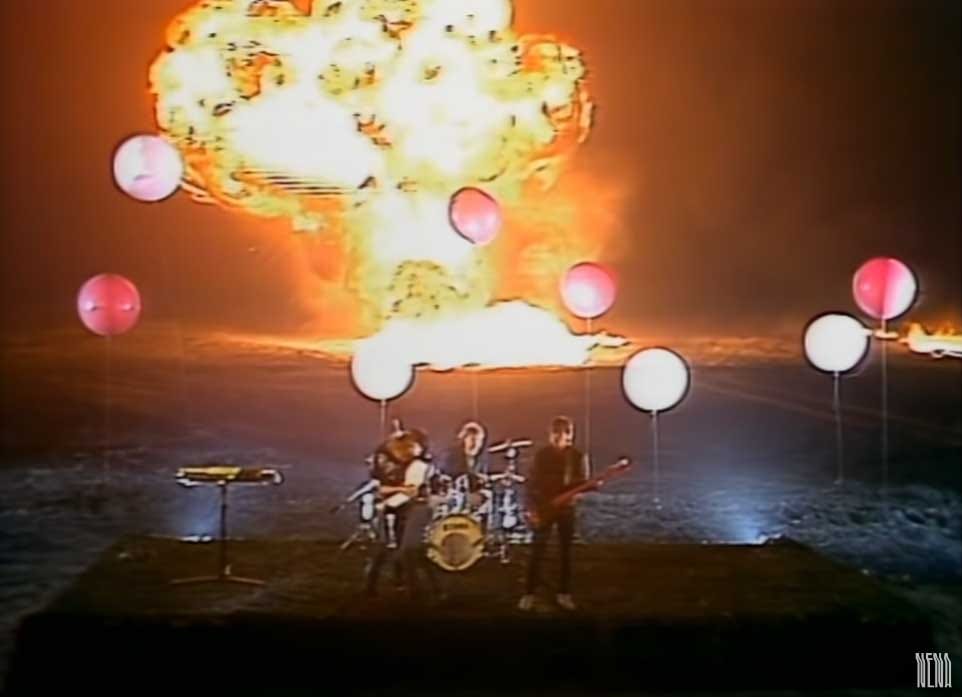 Screenshot of the band with real explosions occurring behind them in the music video for 99 Red Balloons