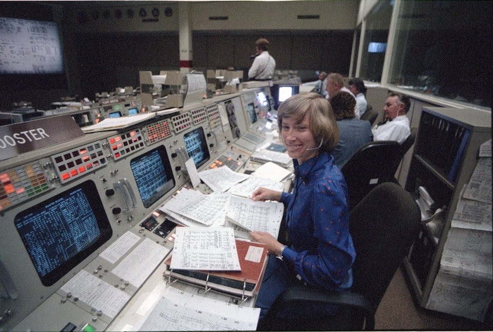 Booster Systems Operator Jenny Howard working at her station. Howard is smiling at the camera while sitting at one of a long line of consoles in a control center, with papers everywhere and colleagues in the background. More rows are in front of her, to the left, with glass viewing windows at right.