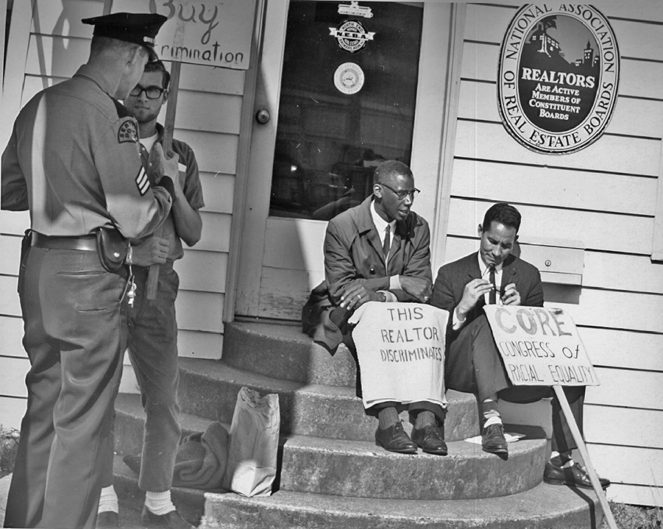 A black and white photo shows two men sitting on the front steps of a building that has light colored siding on it. The sign to the right of the front door reads, “National Association of Real Estate Boards.” The men rest signs at their feet that read, “This Realtor Discriminates” and “CORE. Congress of Racial Equality.” To the left of the steps a protester holds a sign and talks to a police officer that is facing away from the camera.