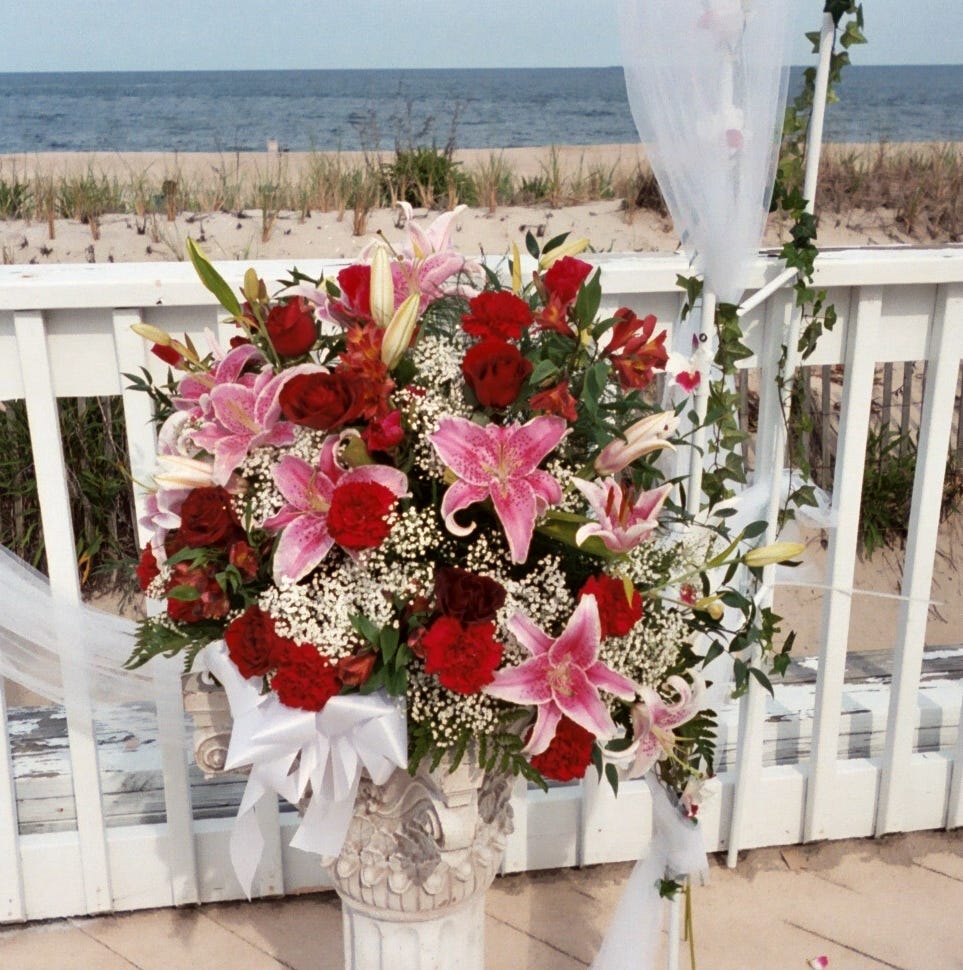 A large basket of flowers including pink stargazers, red roses and babies’ breath. They are on a deck with the ocean visible in on the horizon. A huge white bow anchors the arrangement.