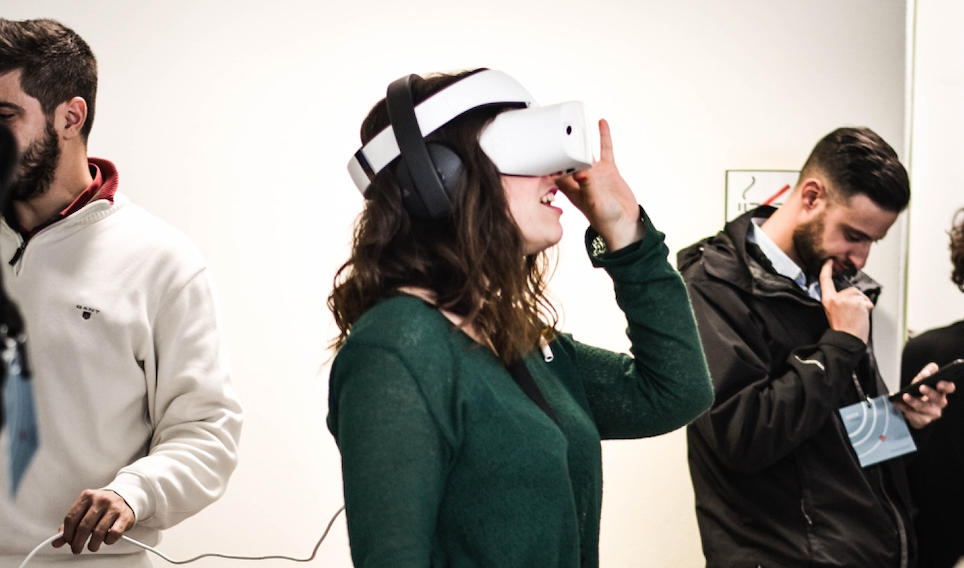 A woman wearing a VR headset. A man is standing next to her reading something and another man holding a cable.