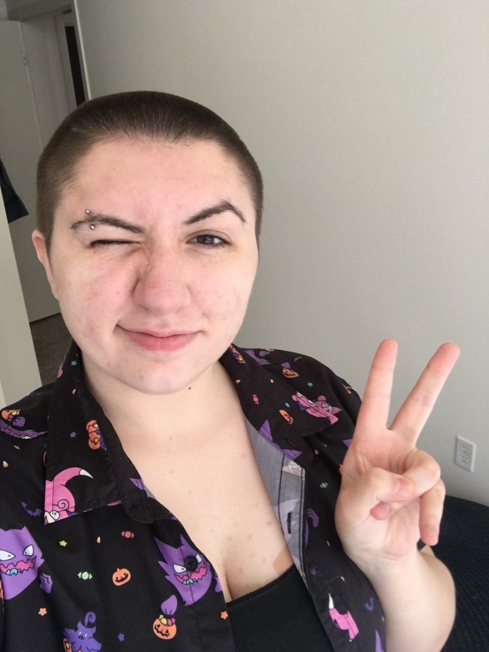 A picture of the author! She’s standing in her room, showing a peace sign to the camera. She winking at the camera and looking forward, smirking. She has an eyebrow piercing over her right eyebrow. She’s wearing a black button up with a pokemon pattern, over a black camisole. Her hair is brown and buzzed short.