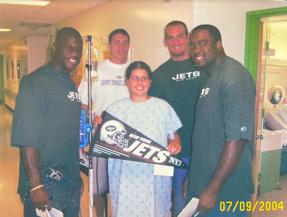 A picture of 14-year-old me after surgery in my hospital gown standing with NY Jets players. I’m holding up a flag that say NY Jets.