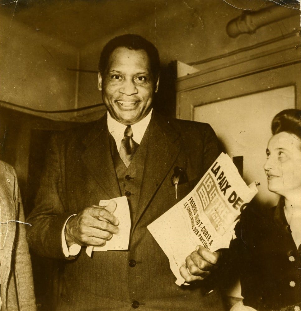 Smiling very tall man wearing a suit and carrying a newspaper. To his right and half cropped from the photograph is a woman smiling and looking upwards towards him