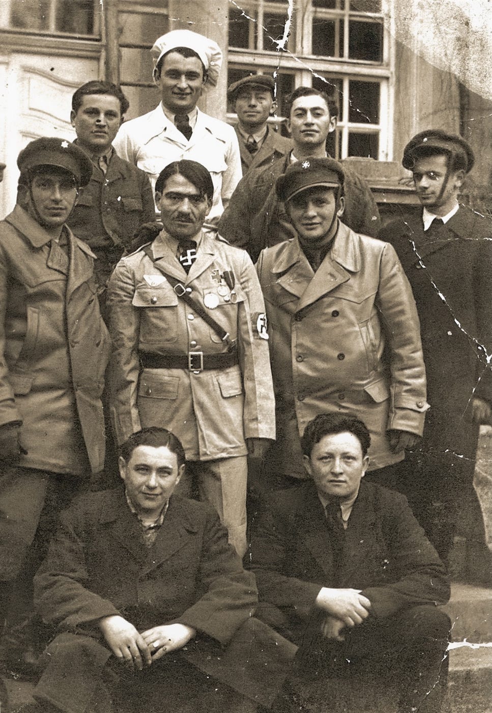 A Jewish displaced person dressed in an Adolf Hitler costume with a belted uniform and swastikas on his necktie and armband stands at the center amid a group of nine other men outside. One of the other men is dressed as a chef and stands behind the man in the Hitler costume.