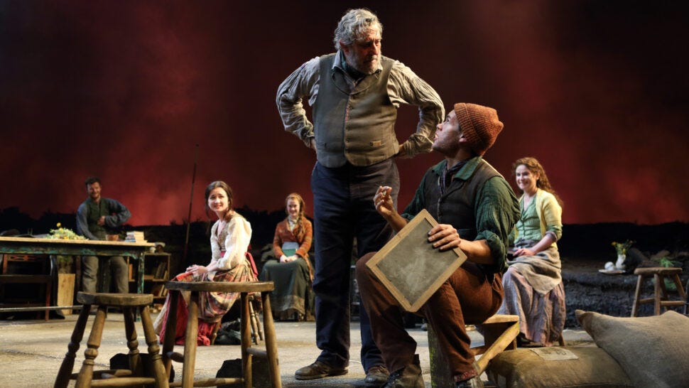 A cast of people in 18th century clothes gathers on a stage in front of a red backdrop