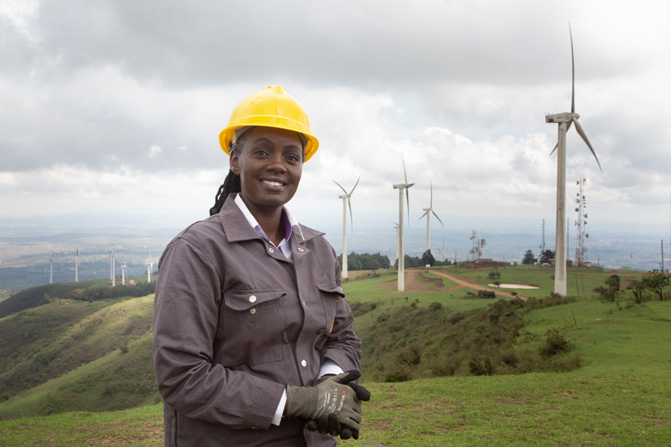 A woman in a yellow hard hat stands in a lush green field filled with windmills.