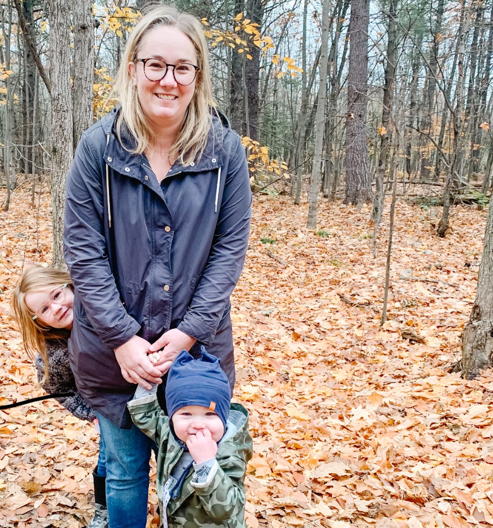 White, blond woman with glasses stands in a forest in autumn with two young children